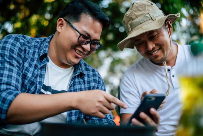 Portrait happy Asian man friends sitting on chair in the camp with talking and looking at smartphone. Cooking set front ground Outdoor cooking, traveling, camping, lifestyle concept.